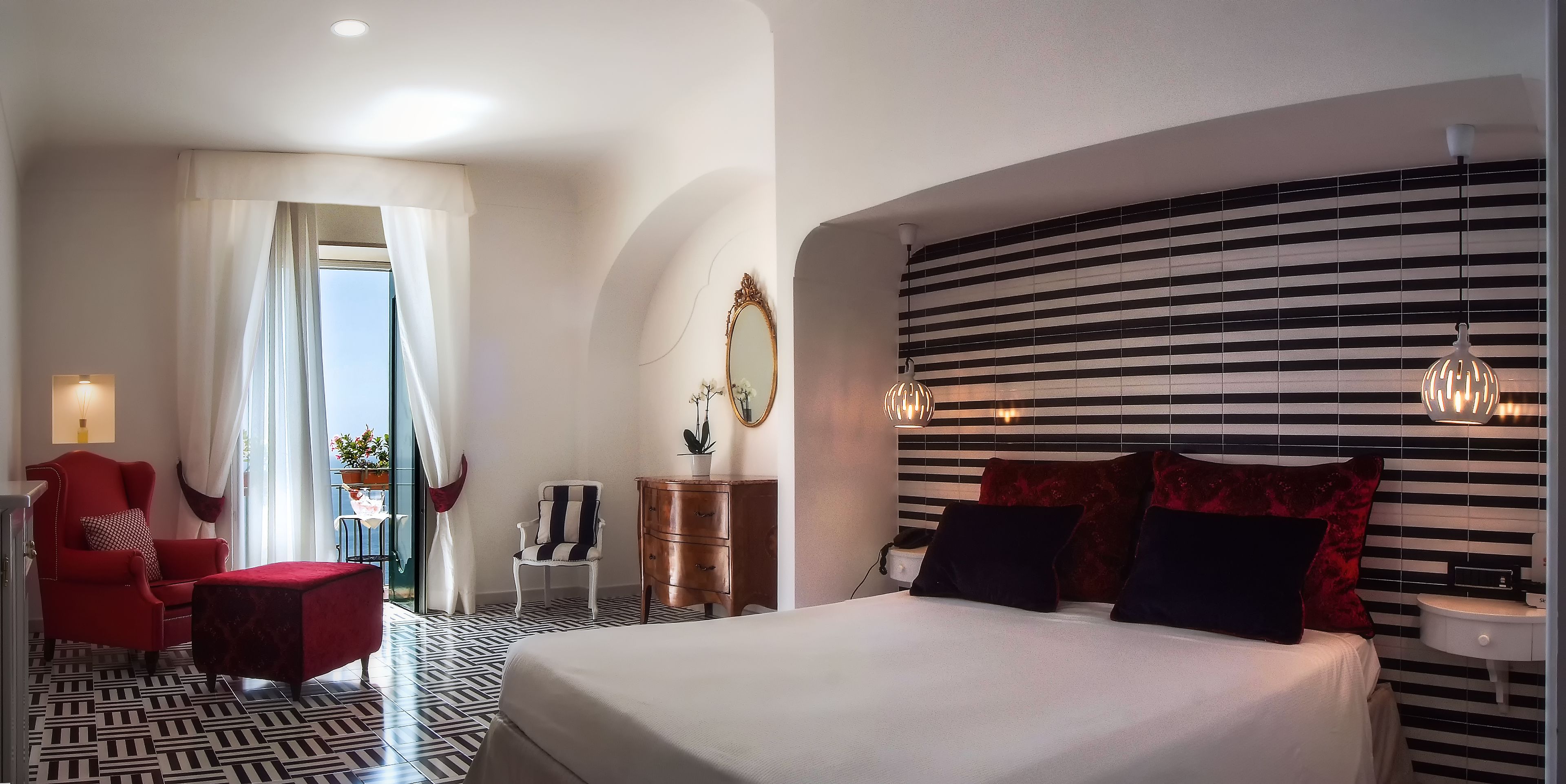 A hotel room with a large bed, a red armchair, a white armchair, a wooden dresser, and a nightstand.
