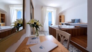 A modern hotel room with a wooden desk, a laptop, a vase with flowers, a large mirror, a bathroom with a sink and vanity, and a view of the sea.