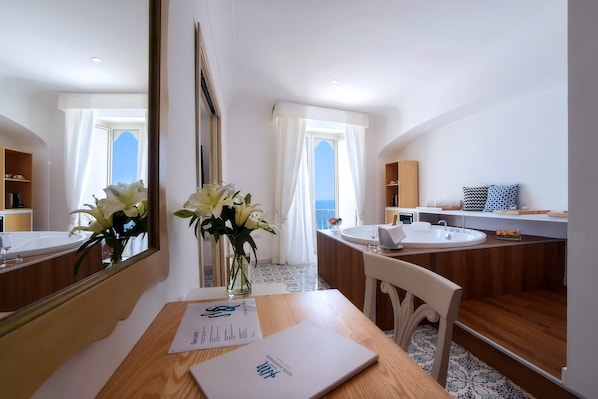 A modern hotel room with a wooden desk, a laptop, a vase with flowers, a large mirror, a bathroom with a sink and vanity, and a view of the sea. - Hotel Montemare (Positano)
