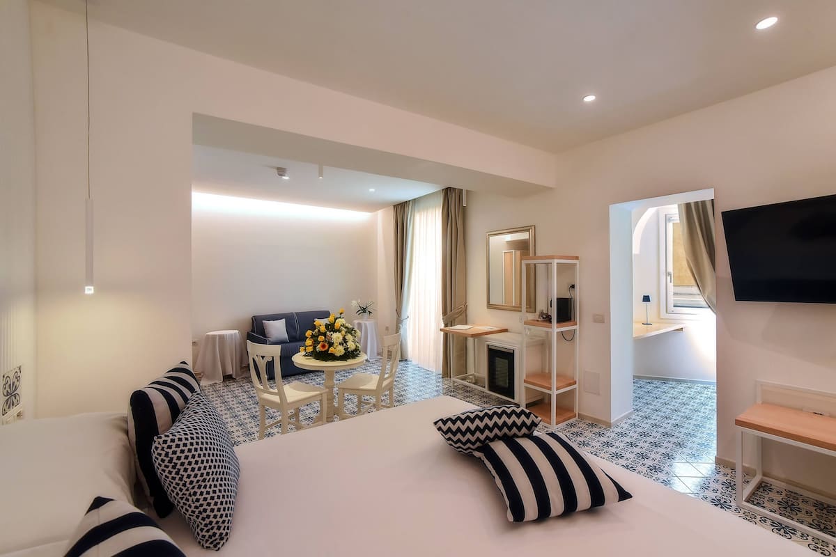 A hotel room with a bed, a desk, a chair, a TV, and a small table with flowers.