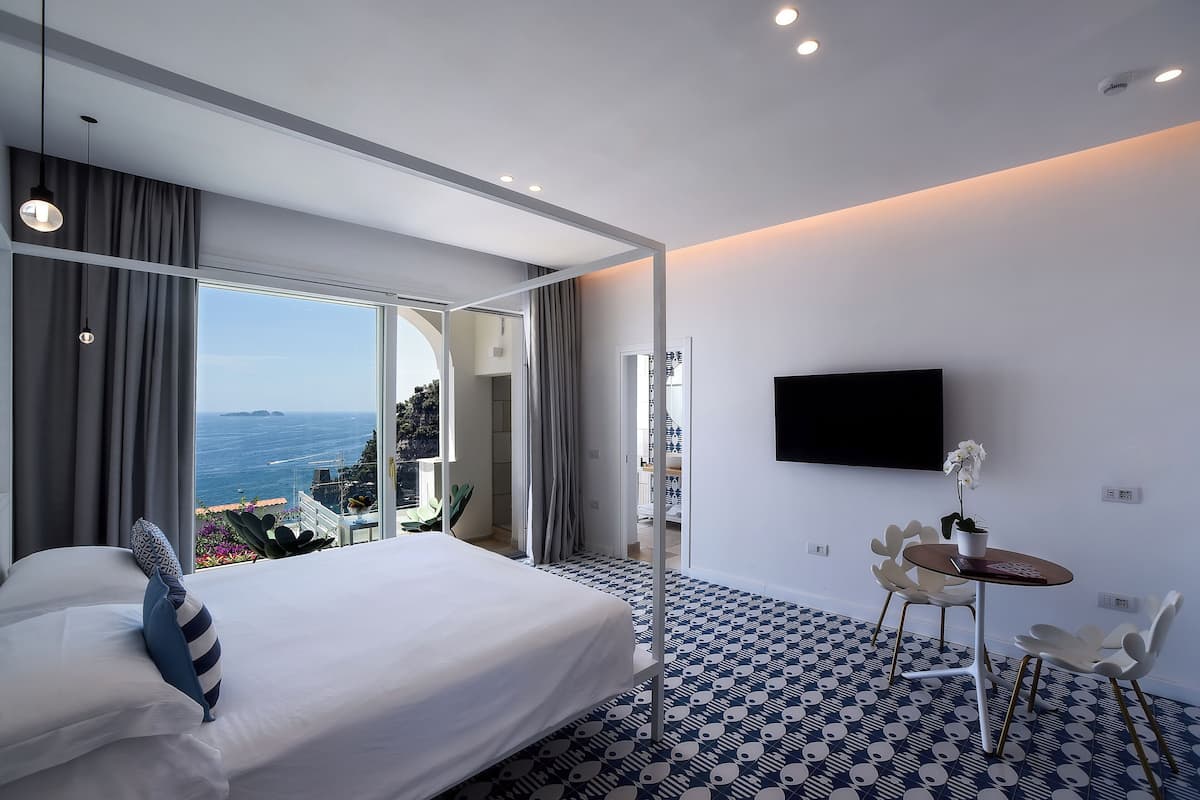 A modern hotel room with a large bed, a TV, a small table with chairs, and a balcony with a view of the sea.