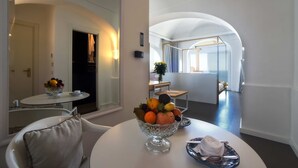 A modern hotel room with a round dining table, a fruit bowl, a tray with a towel, and a view of the adjacent room through an arched doorway.