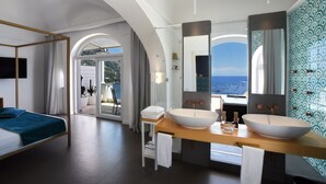 A modern hotel room with a large bed, two sinks, a large mirror, and a view of the sea.