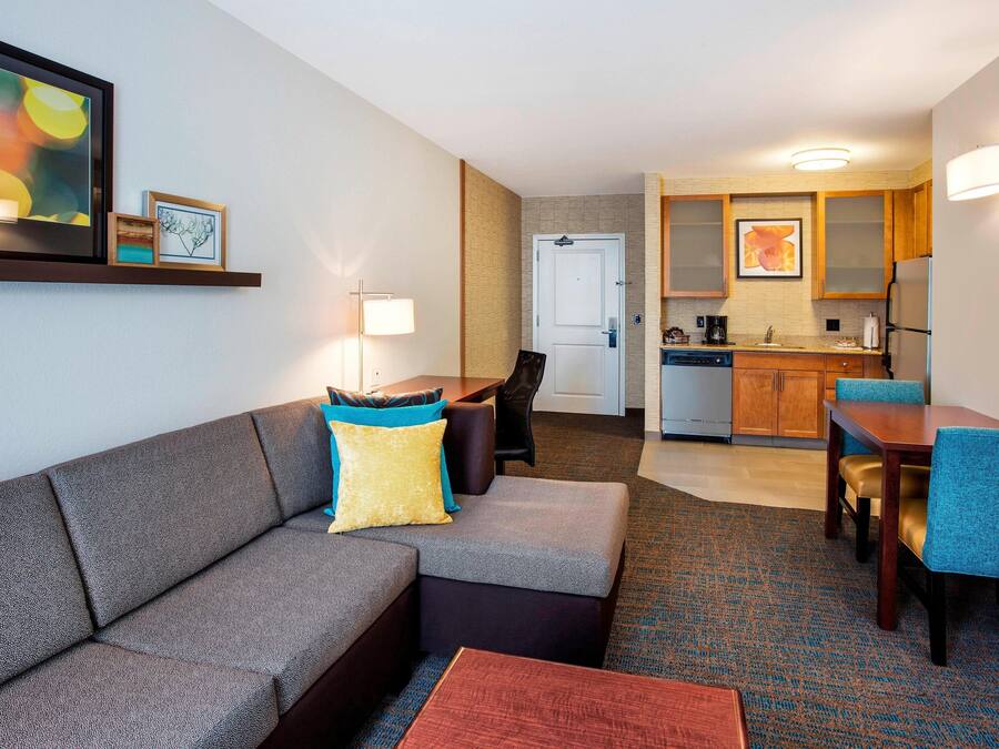 A modern hotel room with a grey sofa, a small table, a dining area with blue chairs, a kitchen area, and a framed artwork on the wall.