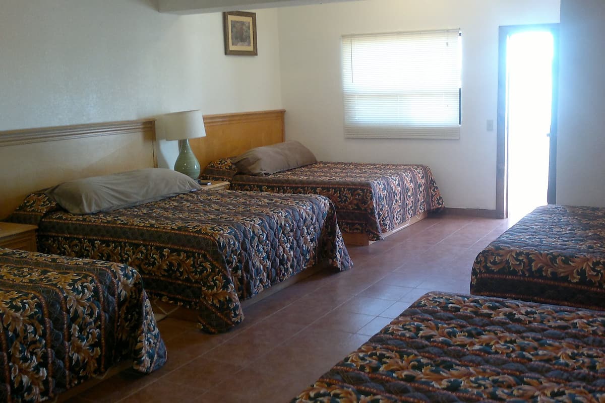 Habitació d'hotel amb dos llits individuals, un doble, una tauleta de nit amb làmpada, una finestra amb persianes i una porta.