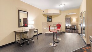 Habitación de hotel con escritorio, dos taburetes rojos, un espejo grande, una cama con cabecero, una mesita de noche con lámpara y un pasillo que conduce a otra habitación.