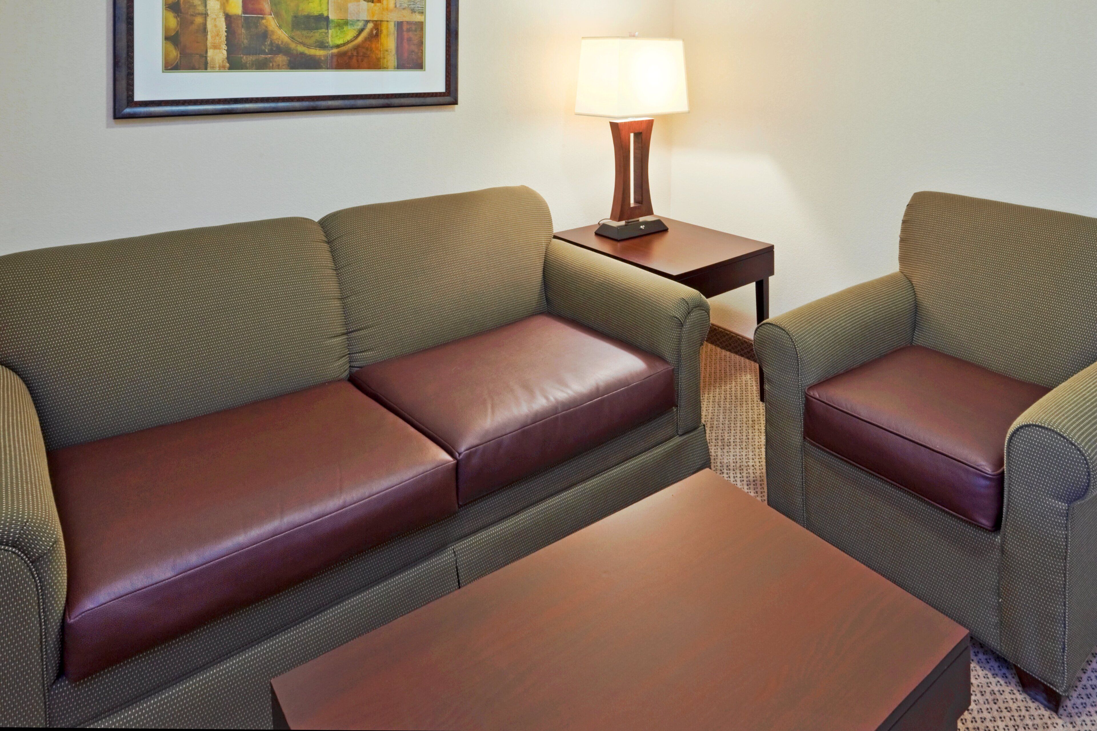A hotel room with a green sofa, a brown leather ottoman, a small wooden side table with a lamp, and a patterned rug.