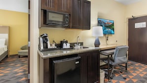 Cocina compacta con microondas, cafetera y lavabo. Incluye un refrigerador pequeño, una silla y un escritorio con lámpara.