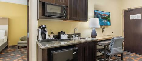 A compact kitchen with a microwave, coffee maker, and sink. There's a small refrigerator, a chair, and a desk with a lamp.