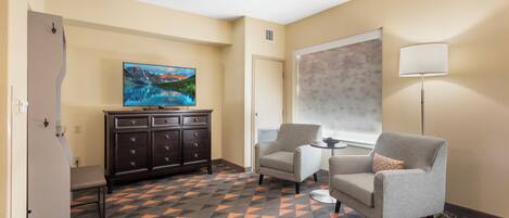 A hotel room with a TV, a desk, two armchairs, a cabinet, and a patterned carpet.