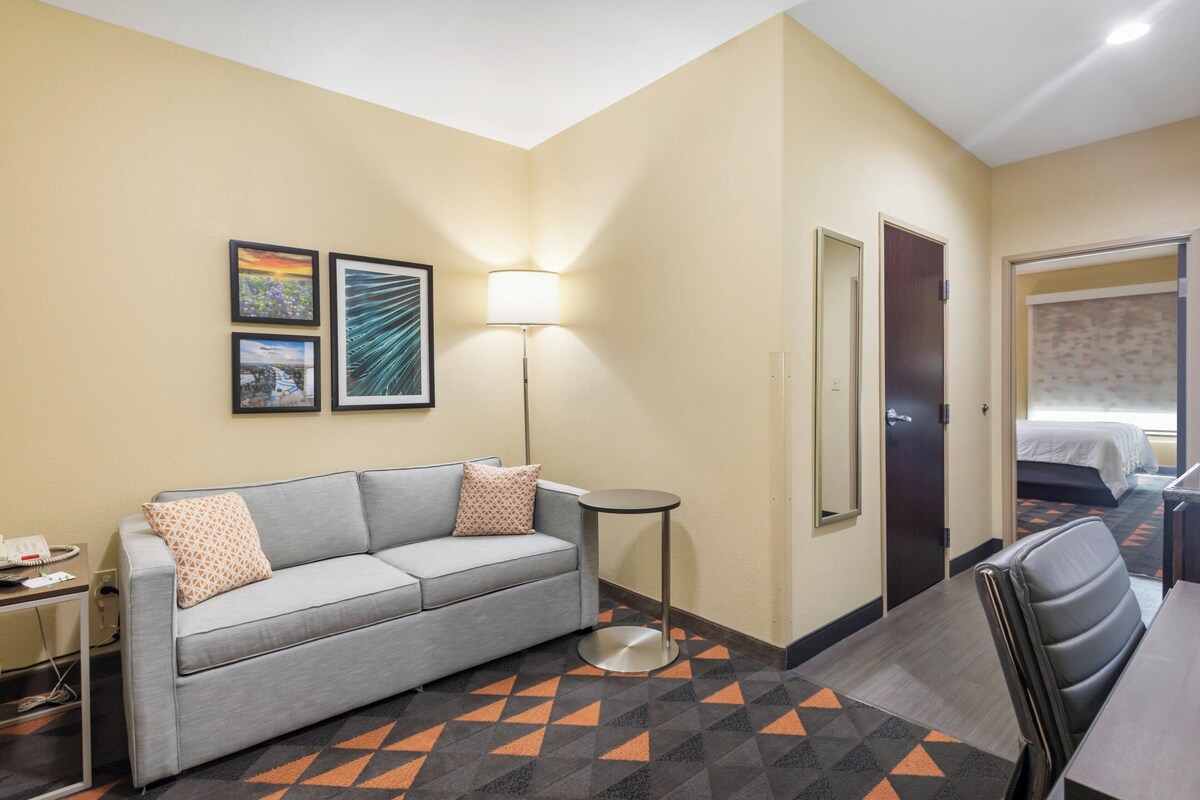 A hotel room with a gray sofa, a small round table, a lamp, and two framed pictures on the wall.