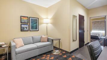 A hotel room with a grey sofa, a small round table, a lamp, and a bed with a patterned bedspread.