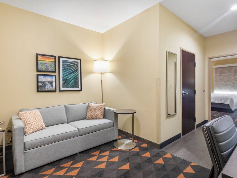 A hotel room with a grey sofa, a small round table, a lamp, and a bed with a patterned bedspread.