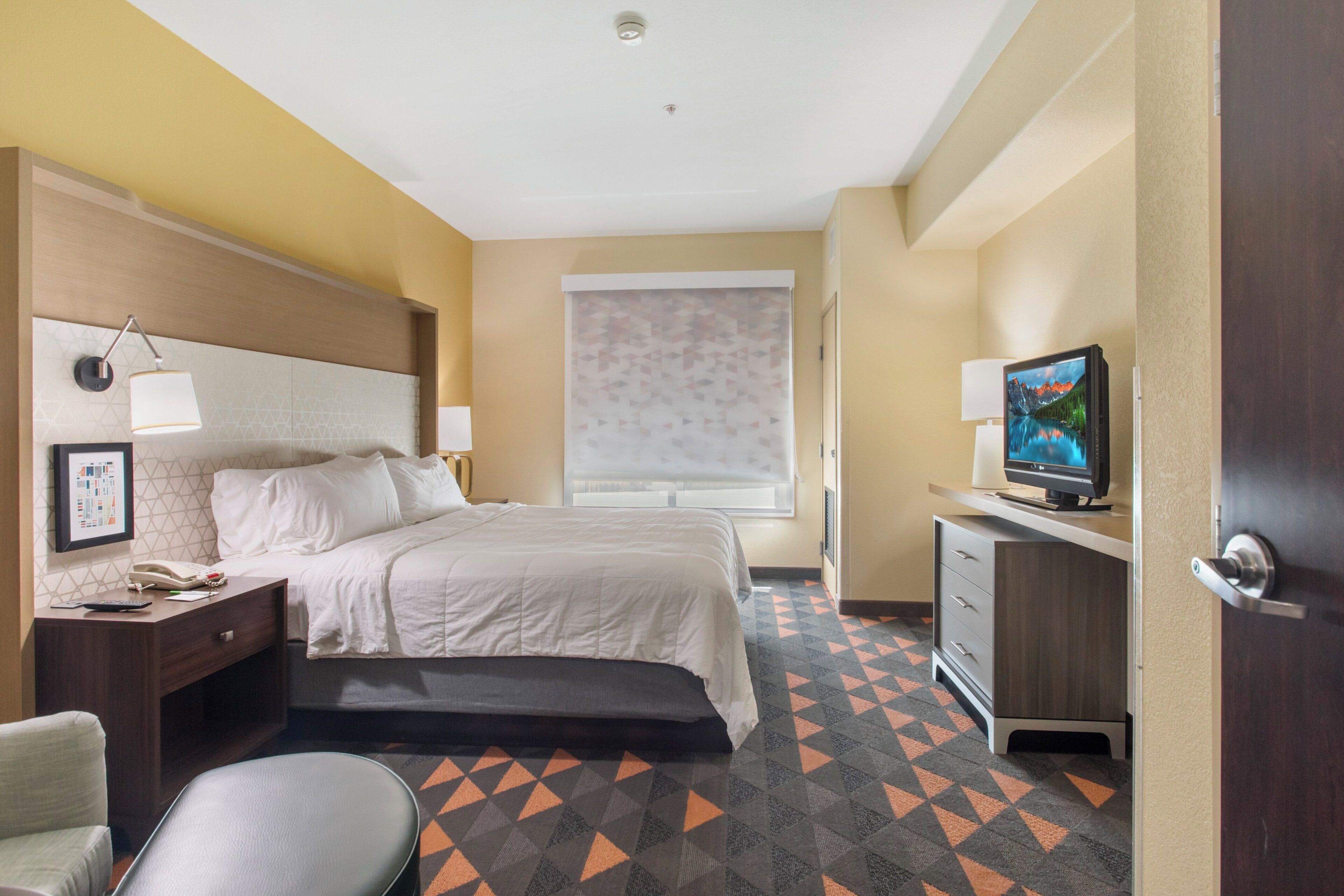 A hotel room with a bed, bedside table, lamp, TV, and a desk with a chair.