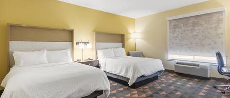 A hotel room with two beds, a large window with a geometric patterned blind, a standing heater, and a chair.