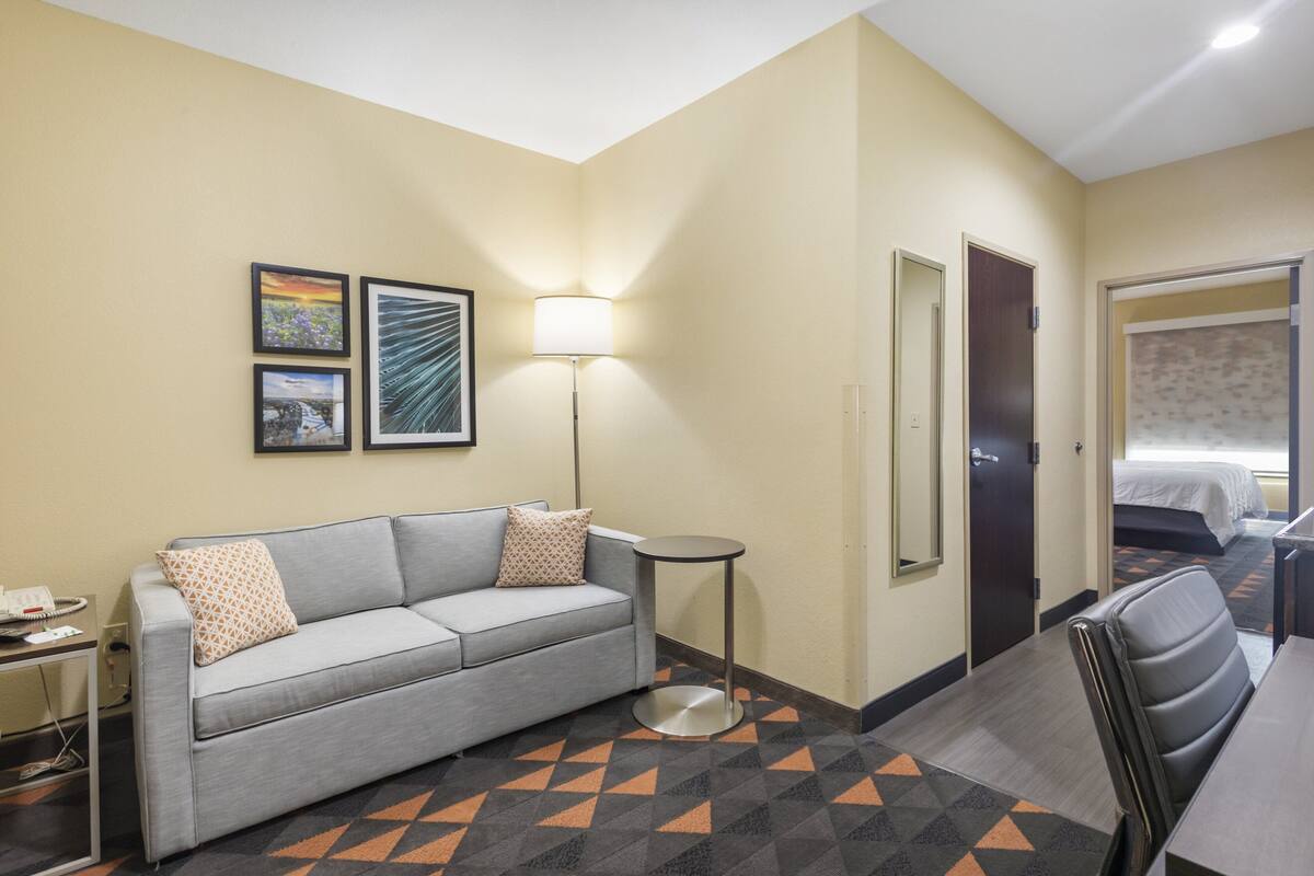 A hotel room with a gray sofa, a small round table, a lamp, and two framed pictures on the wall.