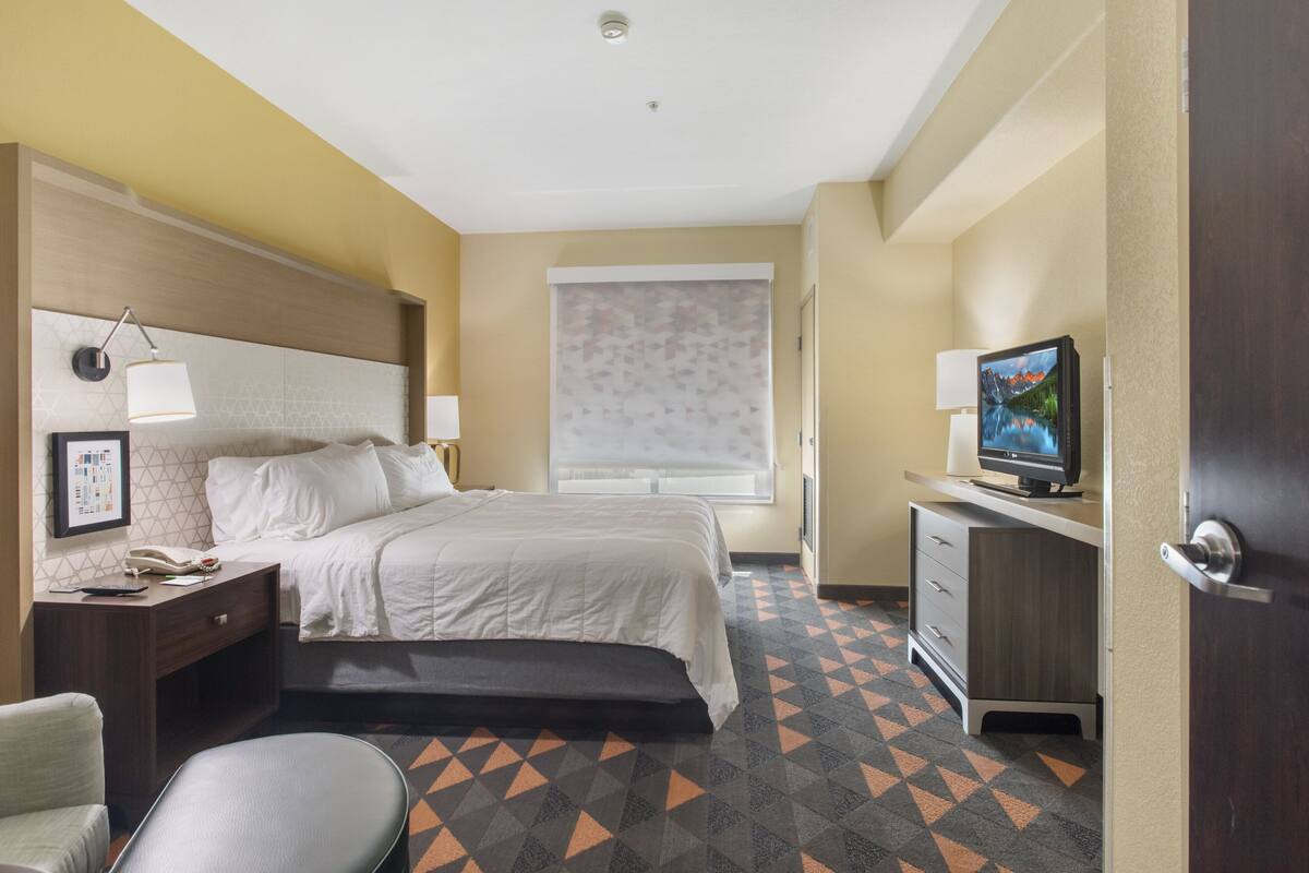 A hotel room with a bed, bedside table, lamp, TV, and a desk with drawers.