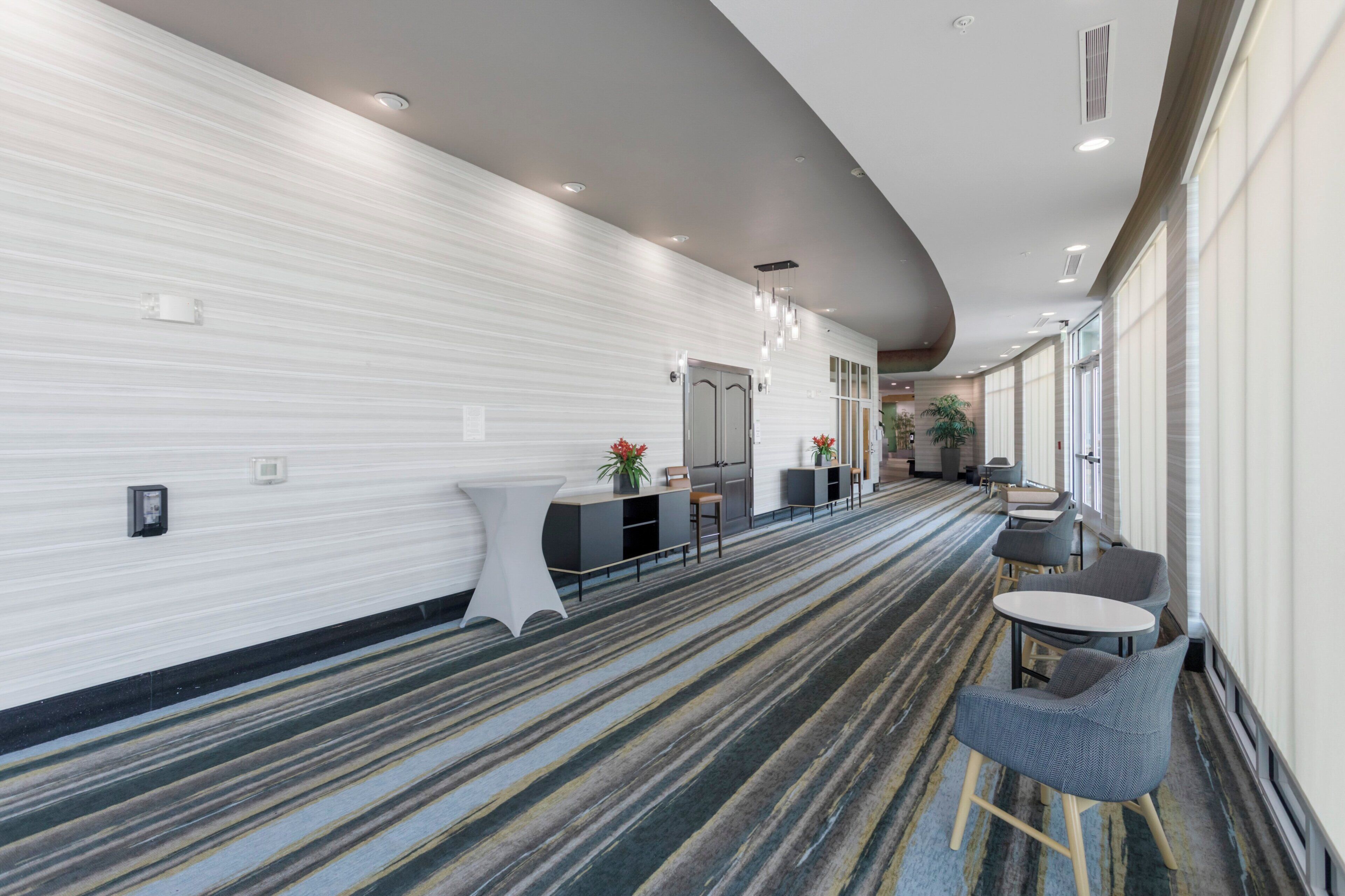 A hotel corridor with a striped carpet, a reception desk, seating area, and a plant.