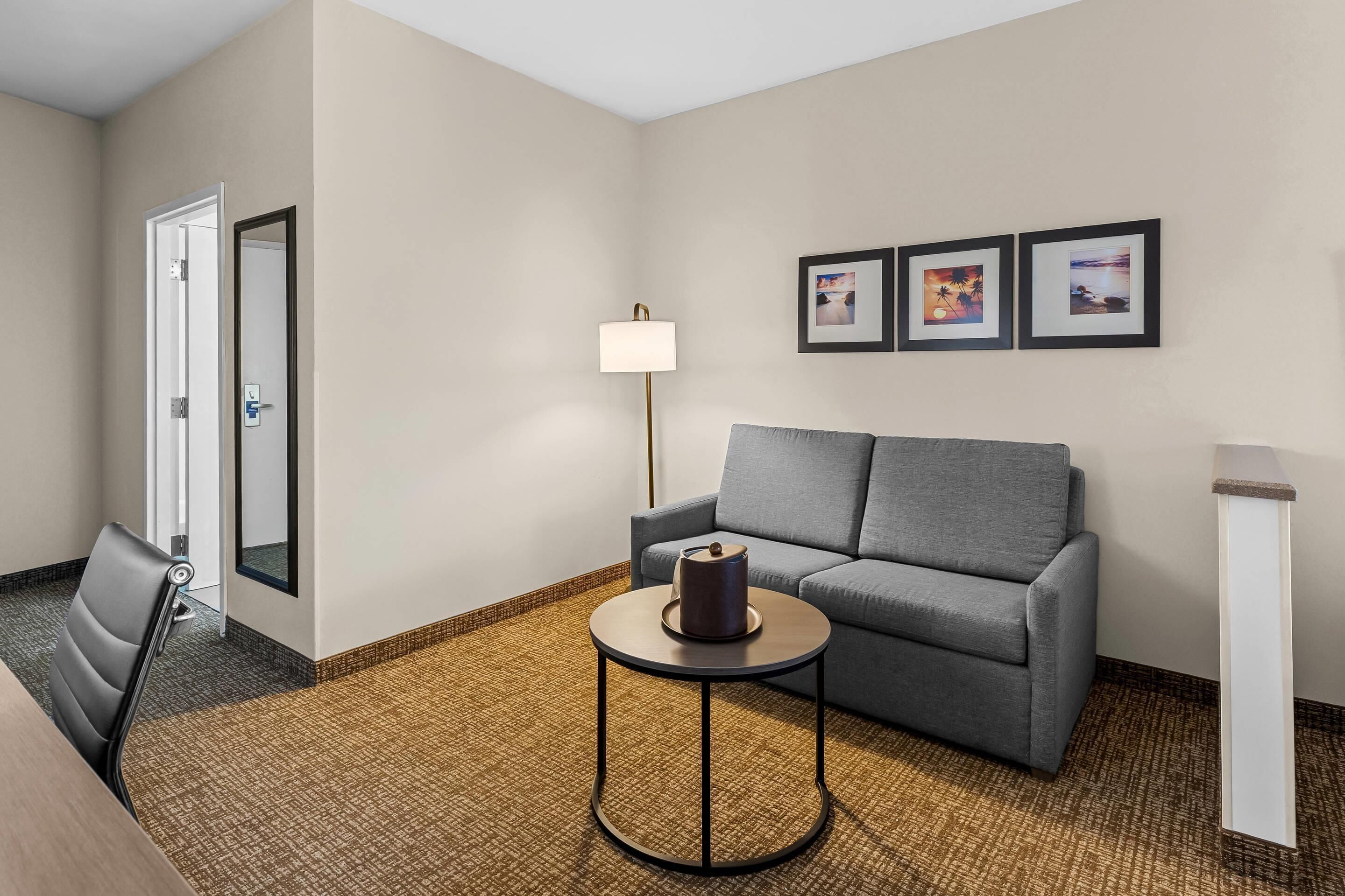 A hotel room with a grey sofa, a small round table with a kettle, a standing lamp, and two framed pictures on the wall.
