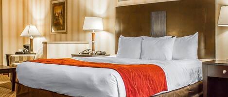 A hotel room with a large bed, two bedside tables, a nightstand, a lamp, a painting, and a patterned carpet.
