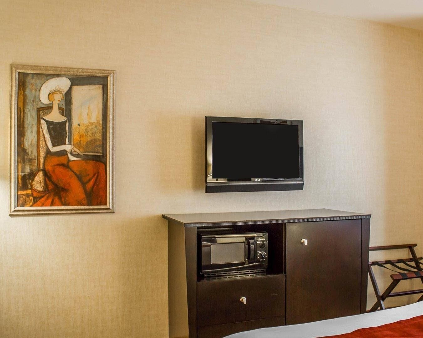 A hotel room with a flat-screen TV mounted on the wall, a painting of a woman, and a dark wooden cabinet with a microwave.