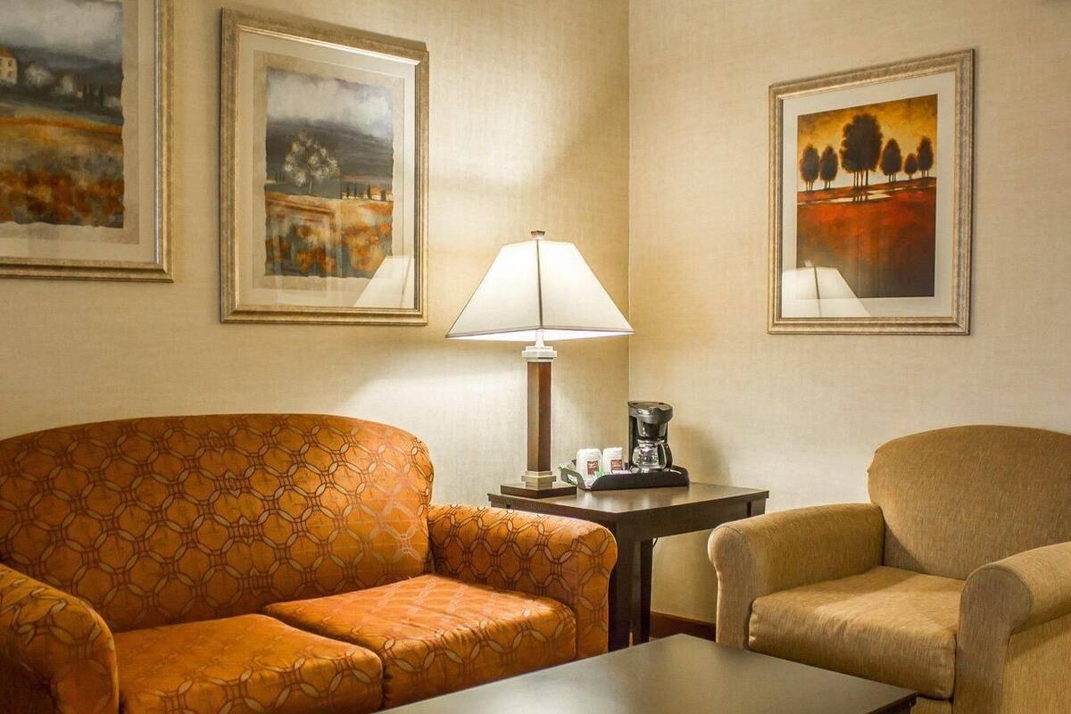A hotel room with a sofa, a chair, a table, a lamp, and two framed paintings on the wall.