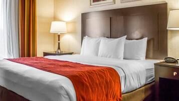 A hotel room with a large bed, a nightstand with a lamp, a clock, and framed artwork on the wall.