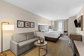 Habitación de hotel con sofá, una pequeña mesa redonda, cama con dos almohadas, televisor montado en la pared y cuadros enmarcados en la pared.