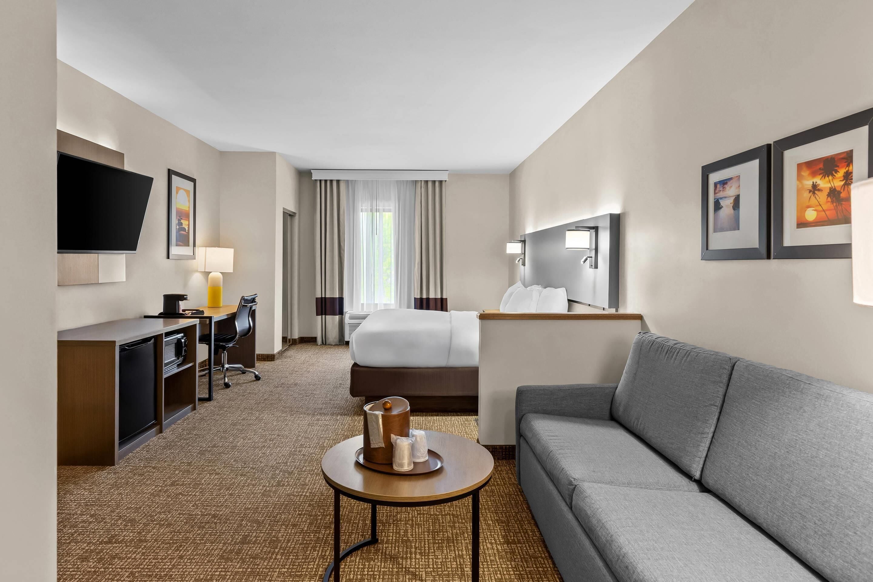 A modern hotel room with a bed, a sofa, a desk with a chair, a TV mounted on the wall, and a small table with a lamp.