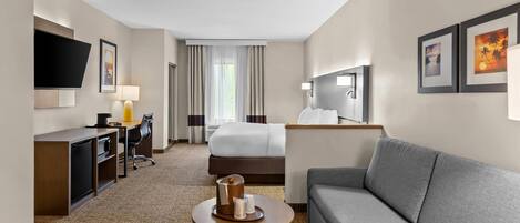 Une chambre d’hôtel moderne équipée d’un lit, d’un canapé, d’un bureau avec une chaise, d’un téléviseur fixé au mur et d’une petite table avec une lampe.
