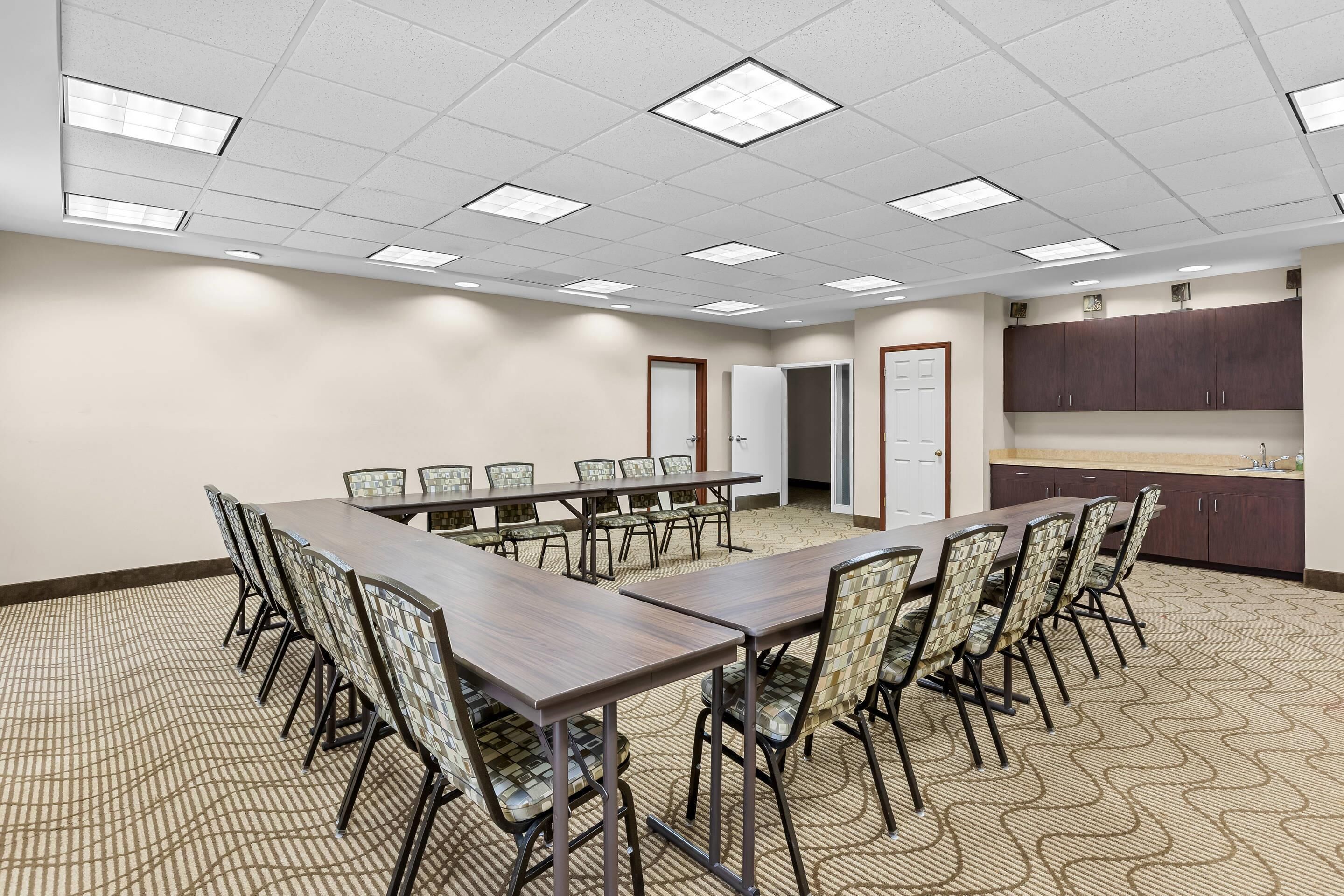 A conference room with a long table, chairs, a kitchenette, and a patterned carpet.