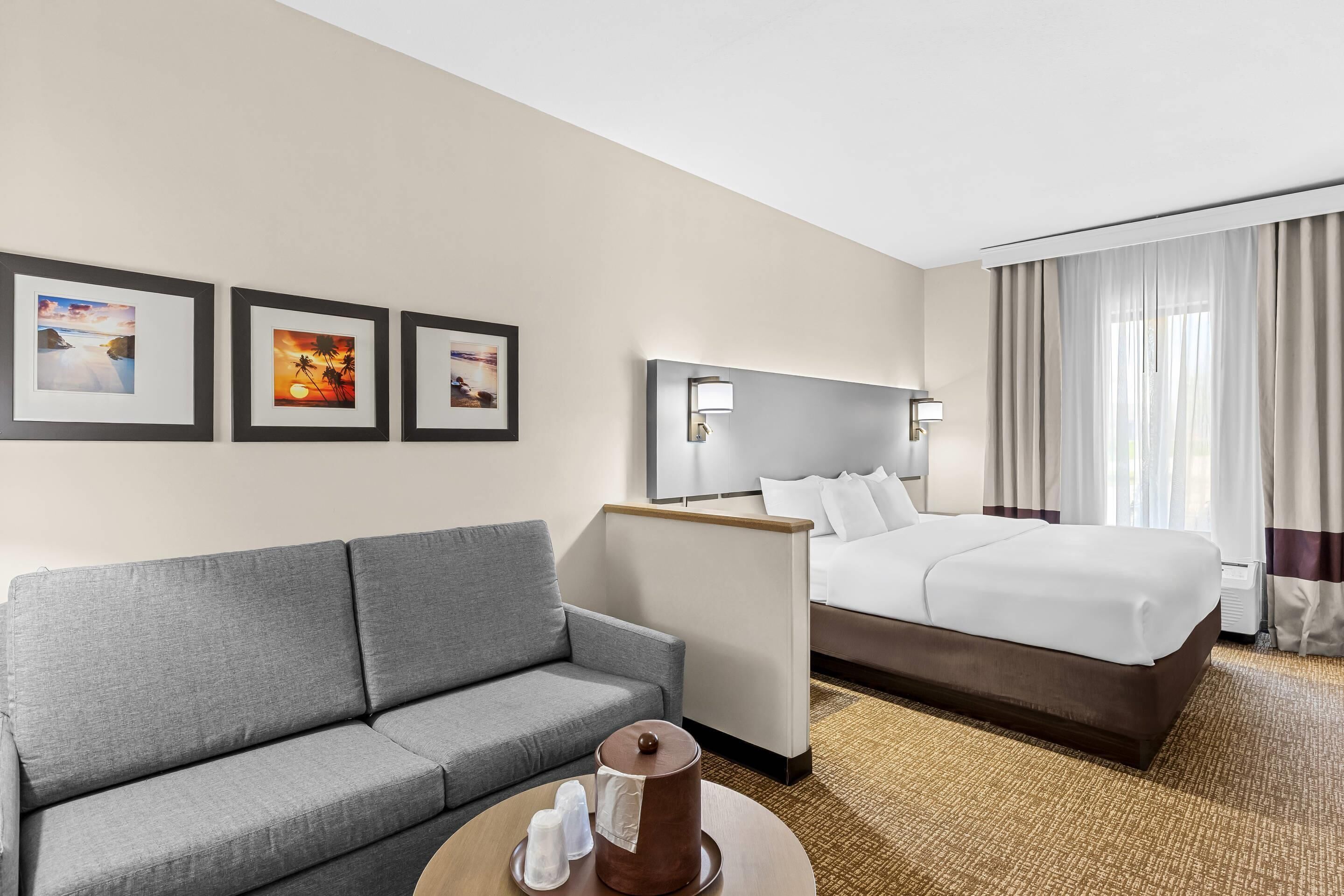 Ein Hotelzimmer mit grauem Sofa, einem Bett mit weißer Bettwäsche, einem runden Tisch mit Lampe und drei gerahmten Bildern an der Wand.
