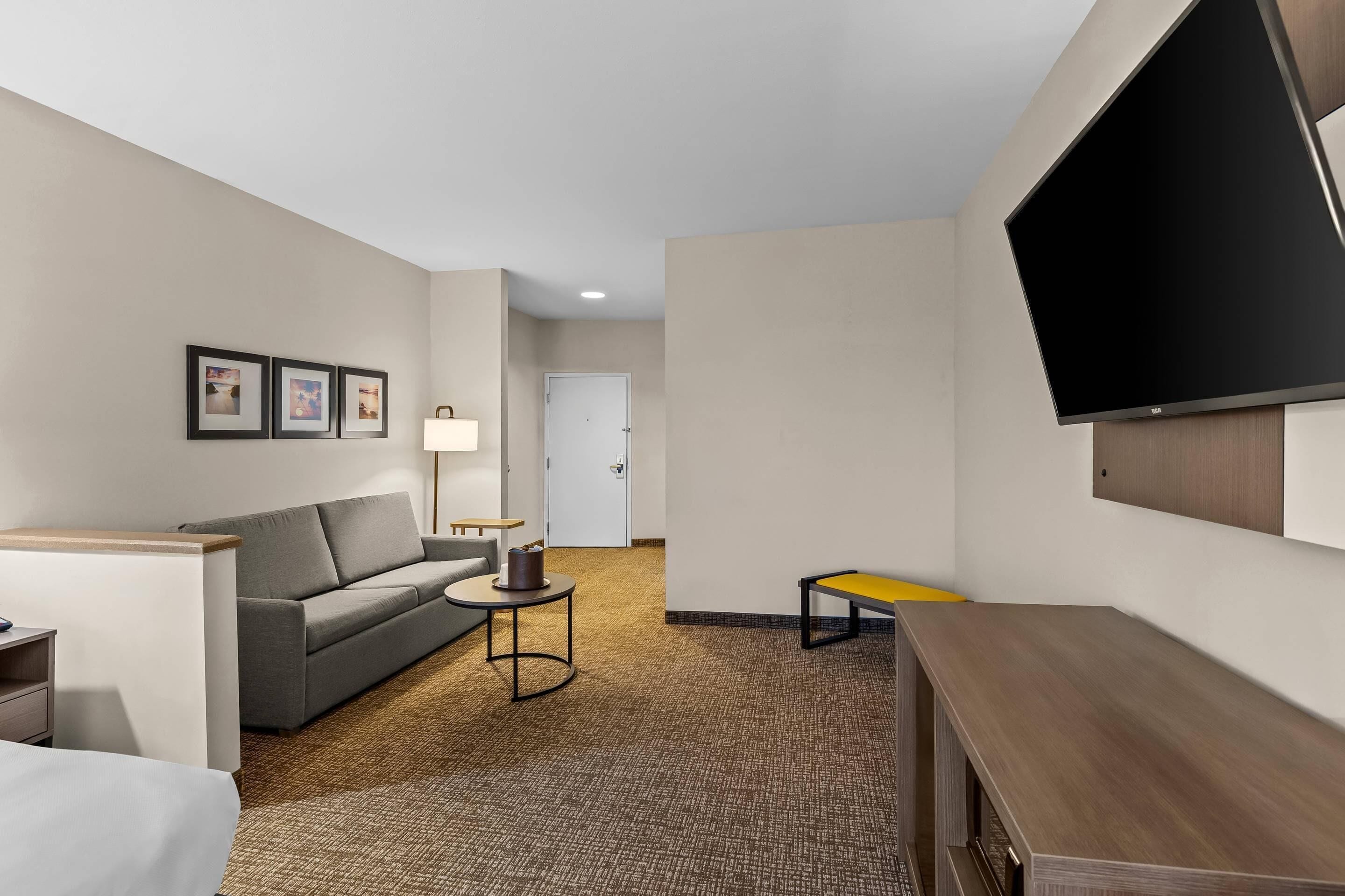 Habitación de hotel con sofá, una pequeña mesa, televisor montado en la pared y una puerta al fondo.