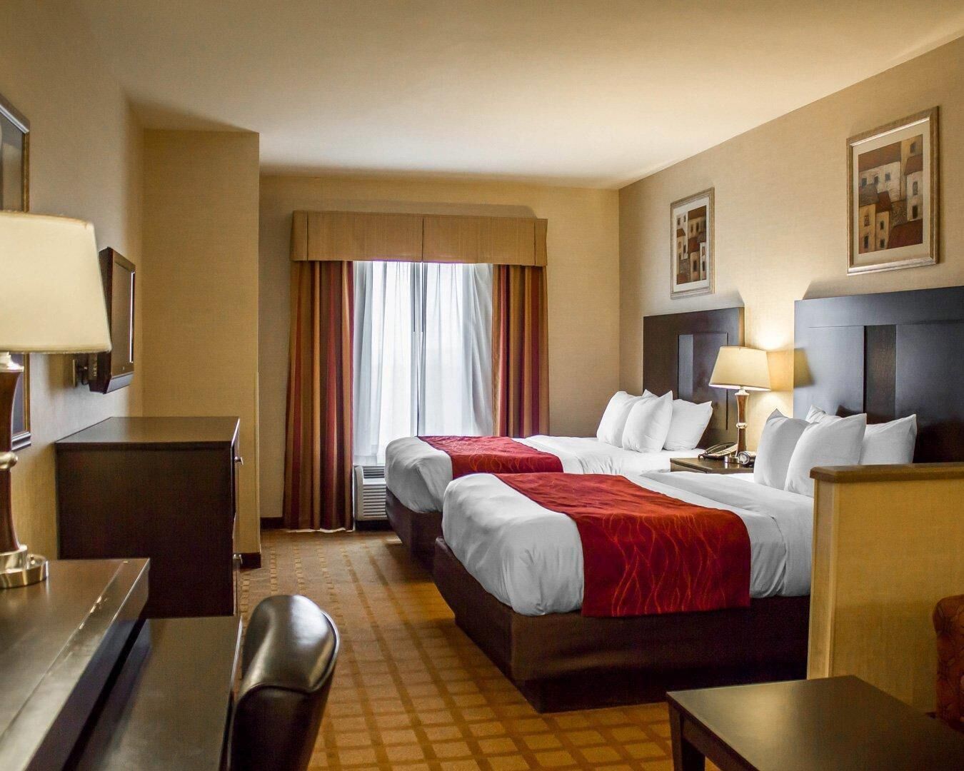 Quarto de hotel com duas camas, uma escrivaninha, uma cadeira, uma mesa de cabeceira e uma janela com cortinas.