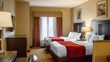 Quarto de hotel com duas camas, uma escrivaninha, uma cadeira, uma mesa de cabeceira e uma janela com cortinas.