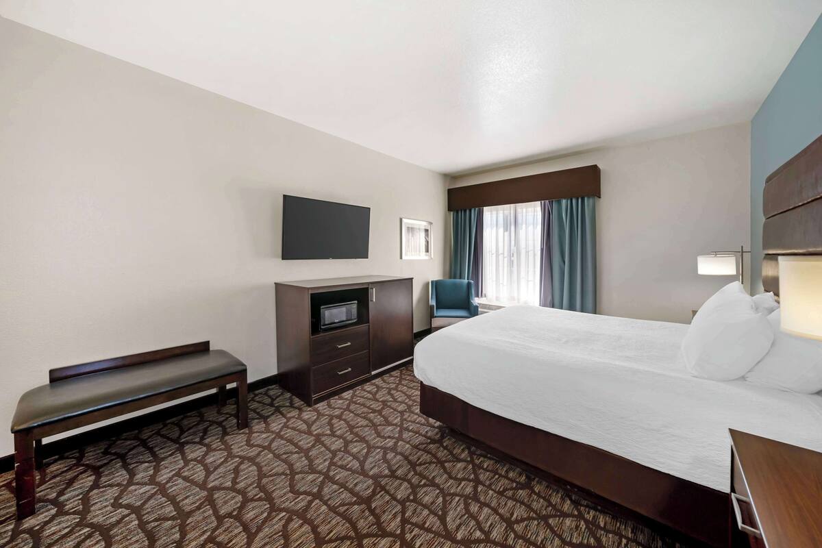 A hotel room with a large bed, a bench, a TV, a desk, and a window with curtains.