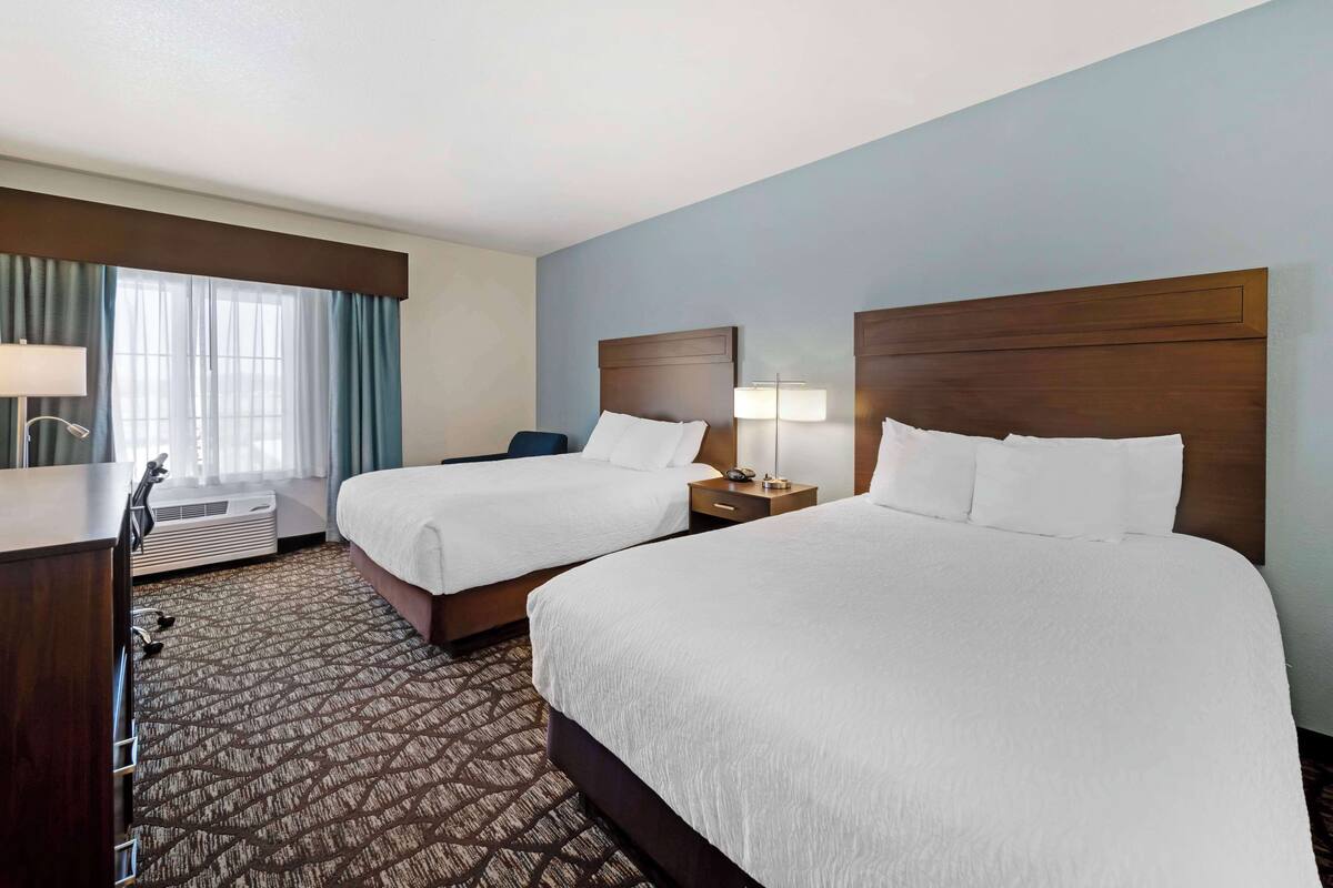 A hotel room with two beds, a desk, a chair, a lamp, and a window with curtains.