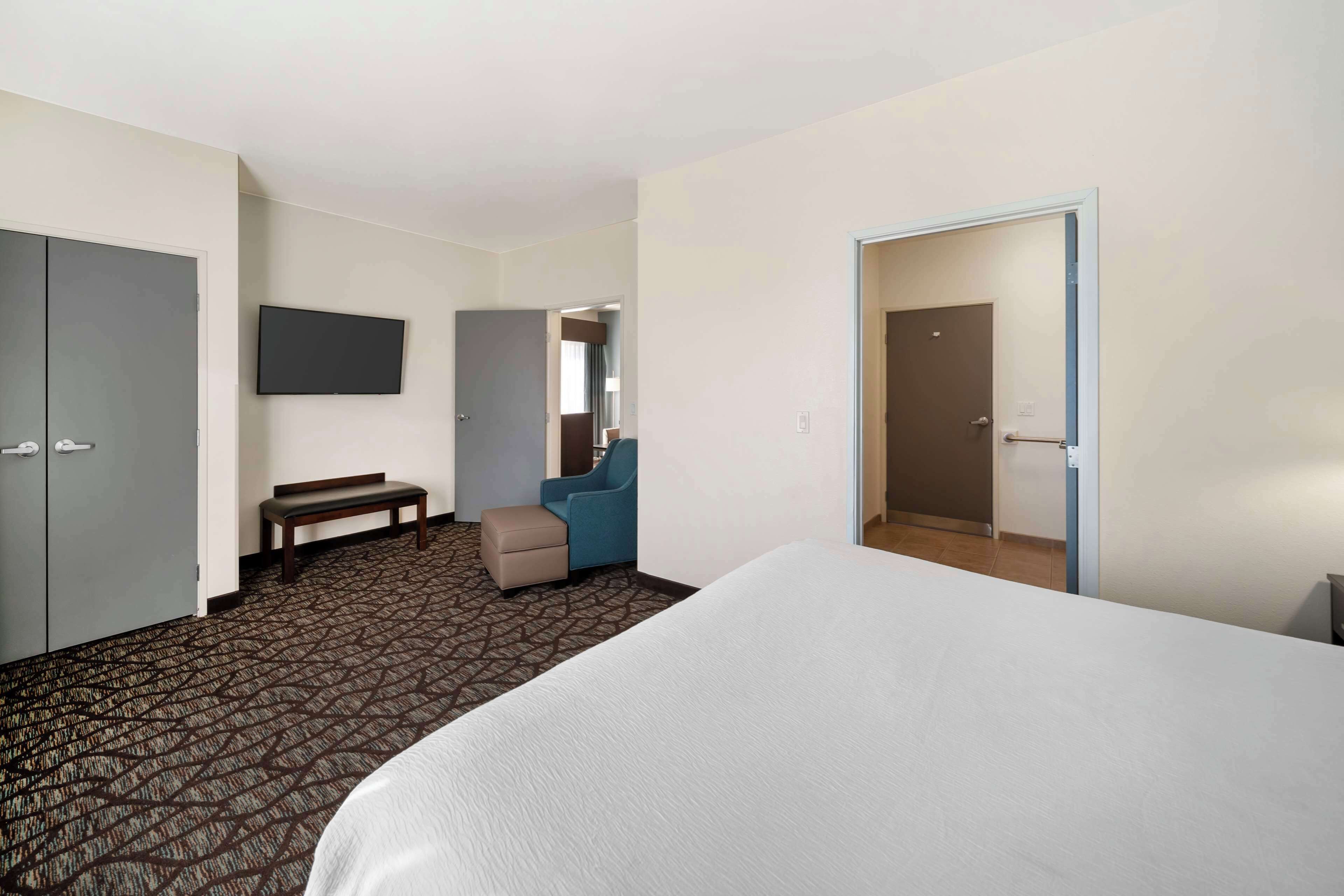 Een hotelkamer met een groot bed, een bureau, een stoel, een bankje, een aan de muur bevestigde televisie en een deur die naar een andere kamer leidt.