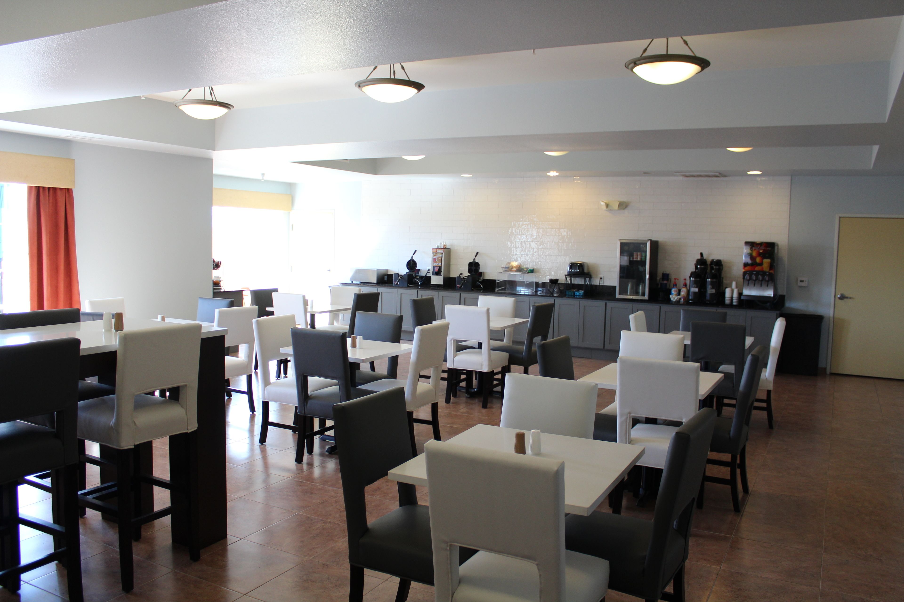 A modern dining area with multiple tables and chairs, a kitchen in the background, and a bright, well-lit space.