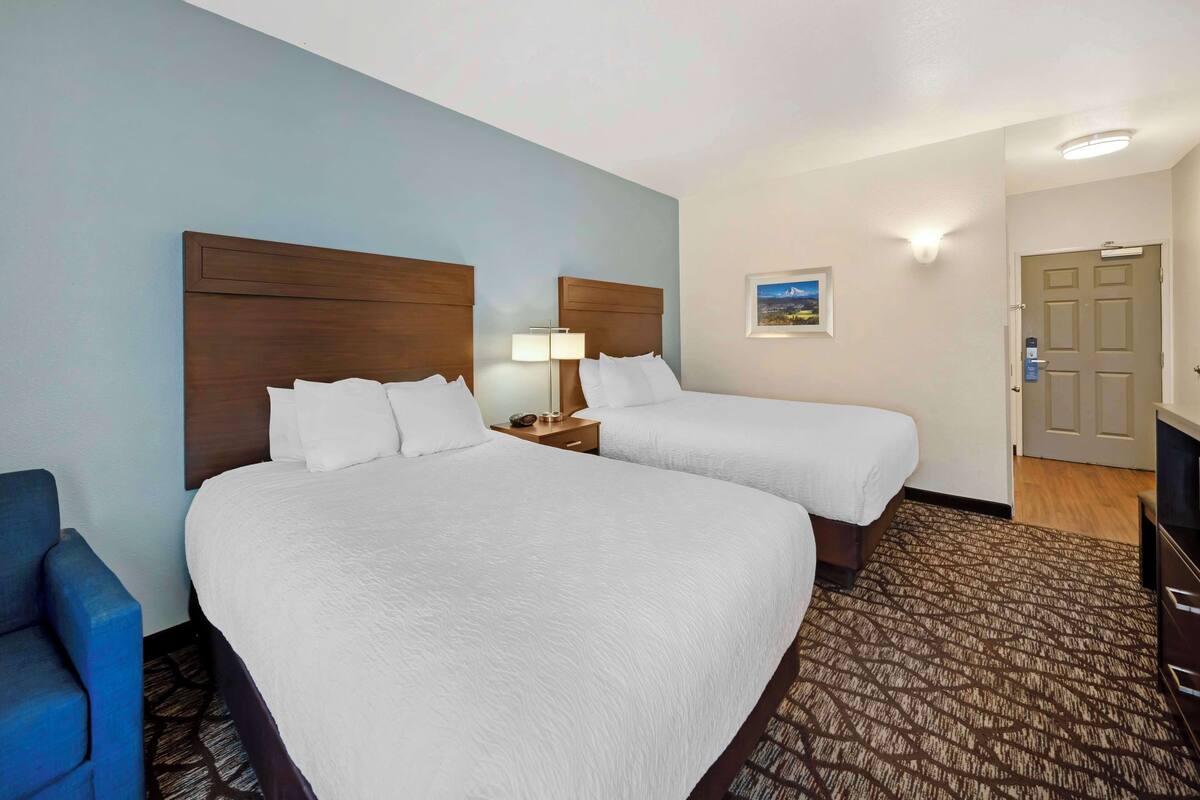 A hotel room with two beds, a desk, a chair, a lamp, and a door.