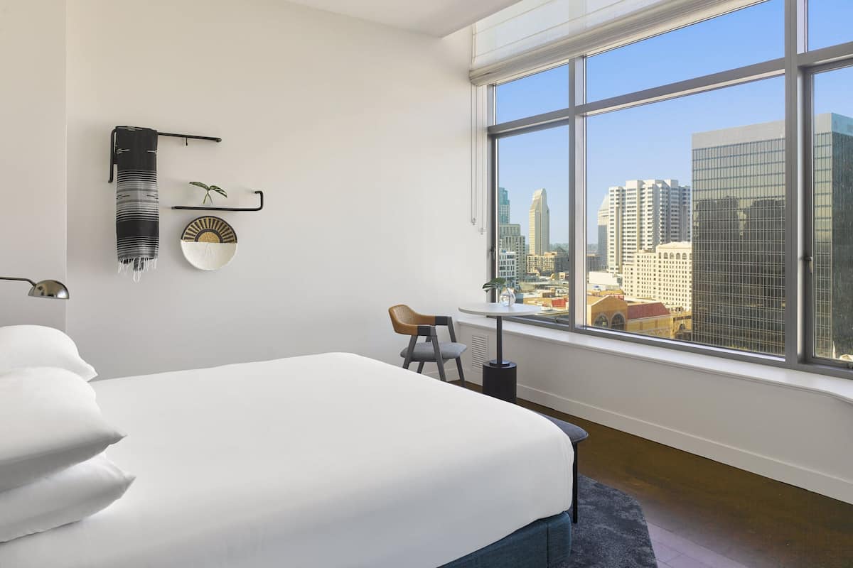 Ein Hotelzimmer mit großem Fenster, einem Bett mit weißer Bettwäsche, einem kleinen Tisch mit Stuhl und einem wandmontierten Regal mit Dekoration.