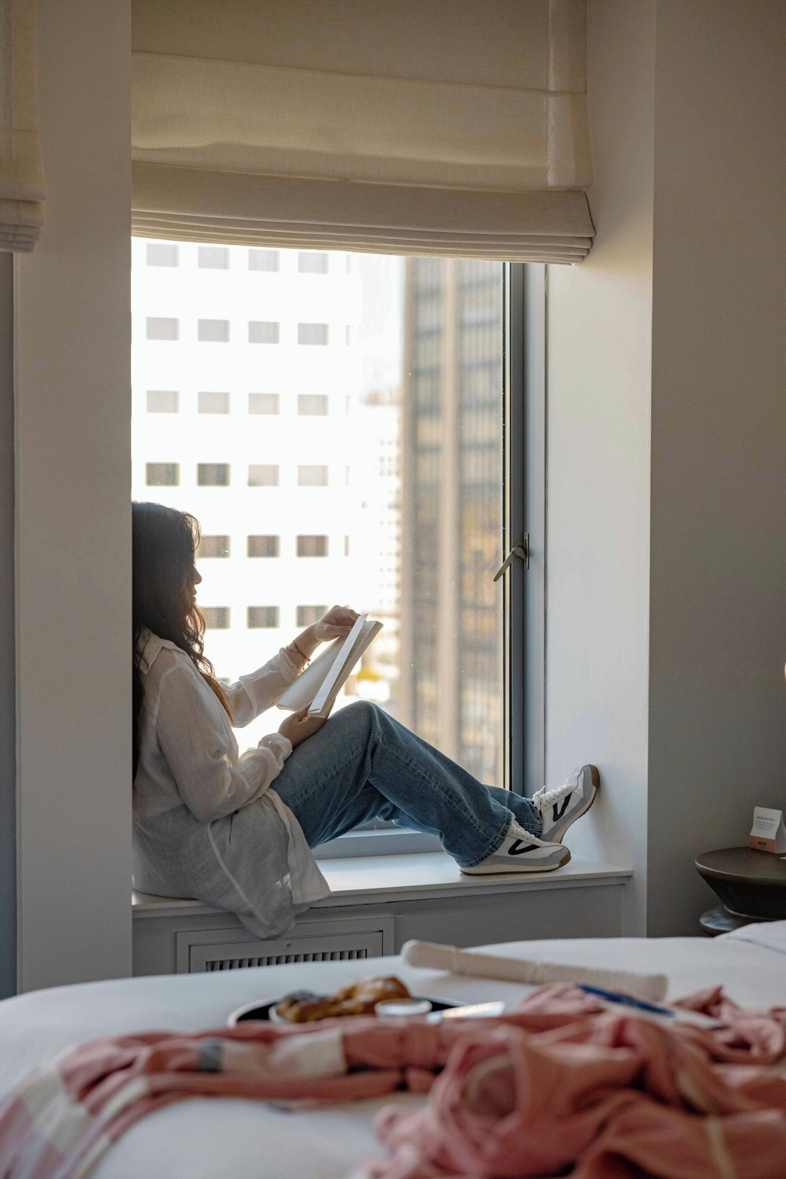 A person sitting by a window in a room with a bed, a small table, and a window blind.