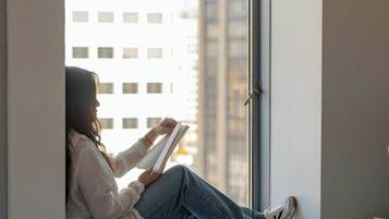 A person sitting by a window in a room with a bed, a small table, and a window blind.