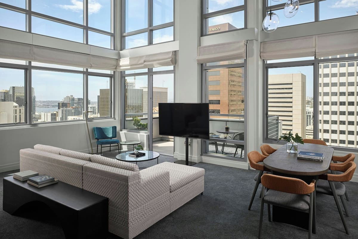 Ein modernes Wohnzimmer mit Sofa, Couchtisch, Fernseher und Essbereich mit Stühlen. Große Fenster bieten Blick auf die Stadt.