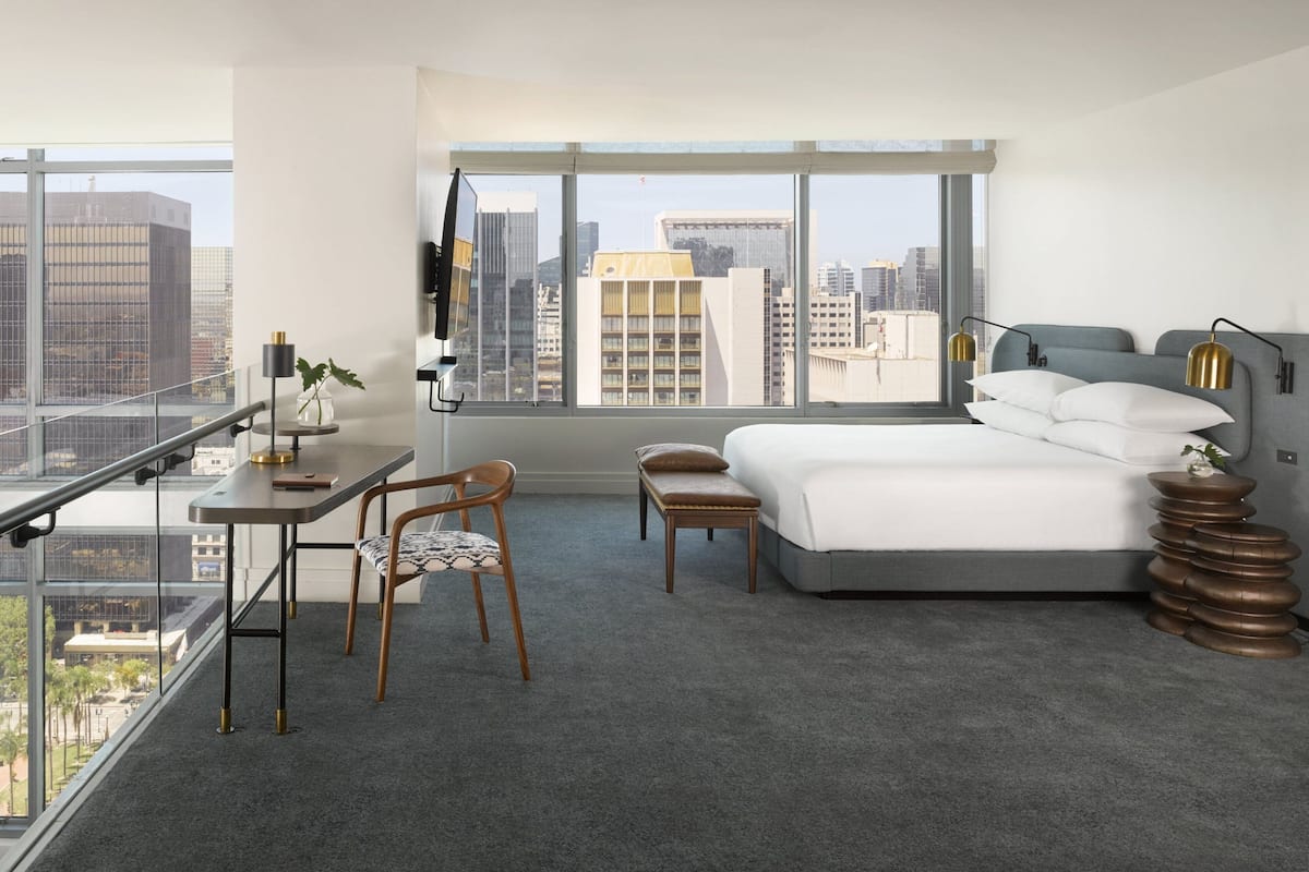 Ein modernes Hotelzimmer mit einem großen Bett, einem Schreibtisch, einem Stuhl und Blick auf die Stadt durch große Fenster.