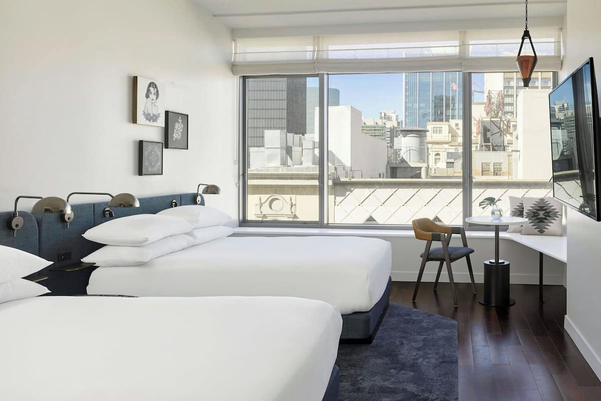 Ein modernes Hotelzimmer mit zwei Betten, einem Schreibtisch und einem großen Fenster mit Blick auf die Stadt.