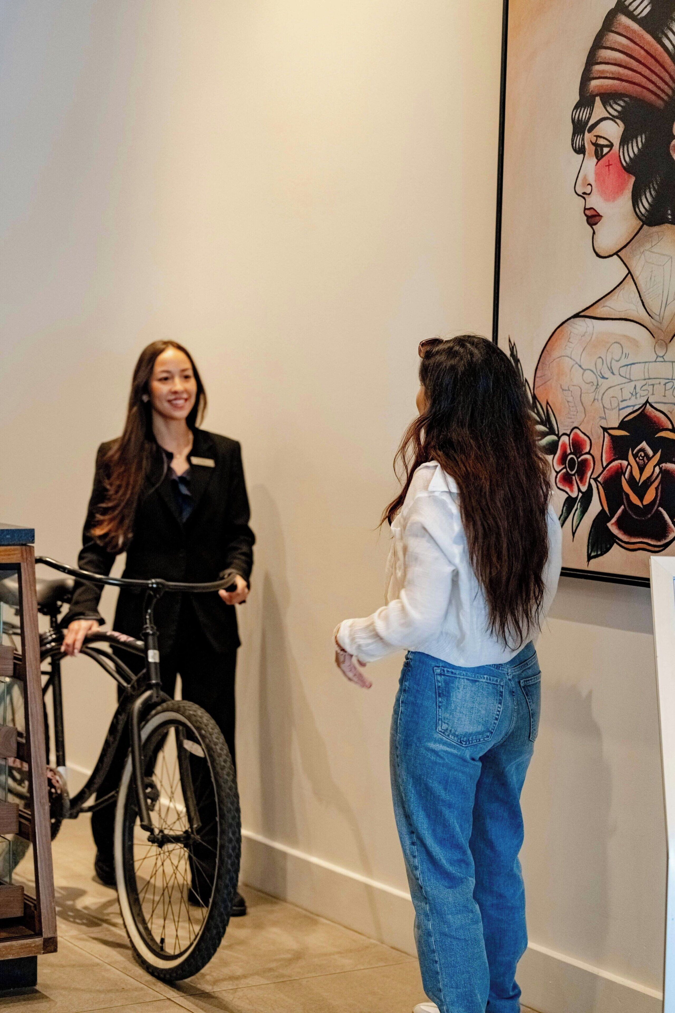 A person on a bicycle, another person standing, and a wall with a large painting.