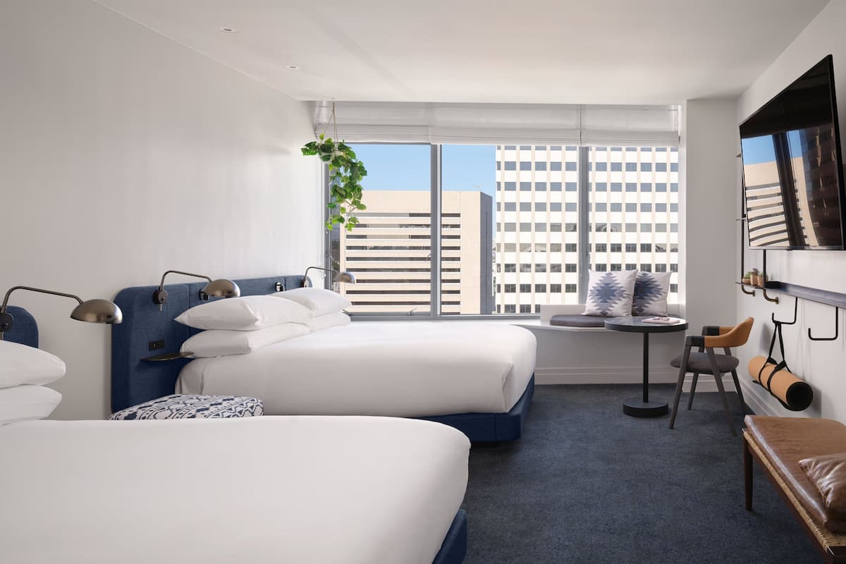Ein modernes Hotelzimmer mit zwei Betten, einem kleinen Tisch, einem Stuhl, einer Couch, einem Fernseher und einem großen Fenster mit Blick auf die Stadt.