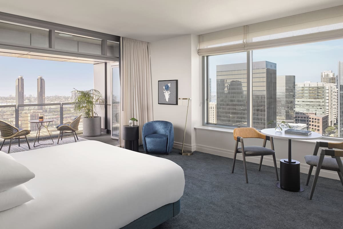 Ein Hotelzimmer mit einem großen Bett, einem Schreibtisch mit zwei Stühlen, Blick auf die Stadt und einem Balkon mit Korbstühlen und einem Blumentopf.