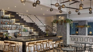 A modern bar area with a bar counter, stools, and tables. There are shelves with bottles and glasses, and a staircase leading to another level. - Alma San Diego Downtown, a Tribute Portfolio Hotel (San Diego)